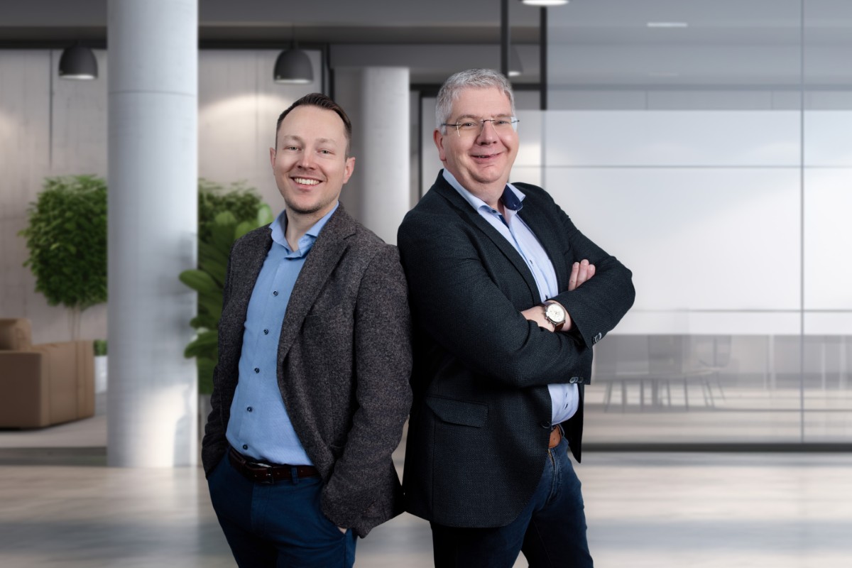 Zwei Männer in Sakkos stehen in einem modernen Büro, lächeln und sehen selbstbewusst aus.